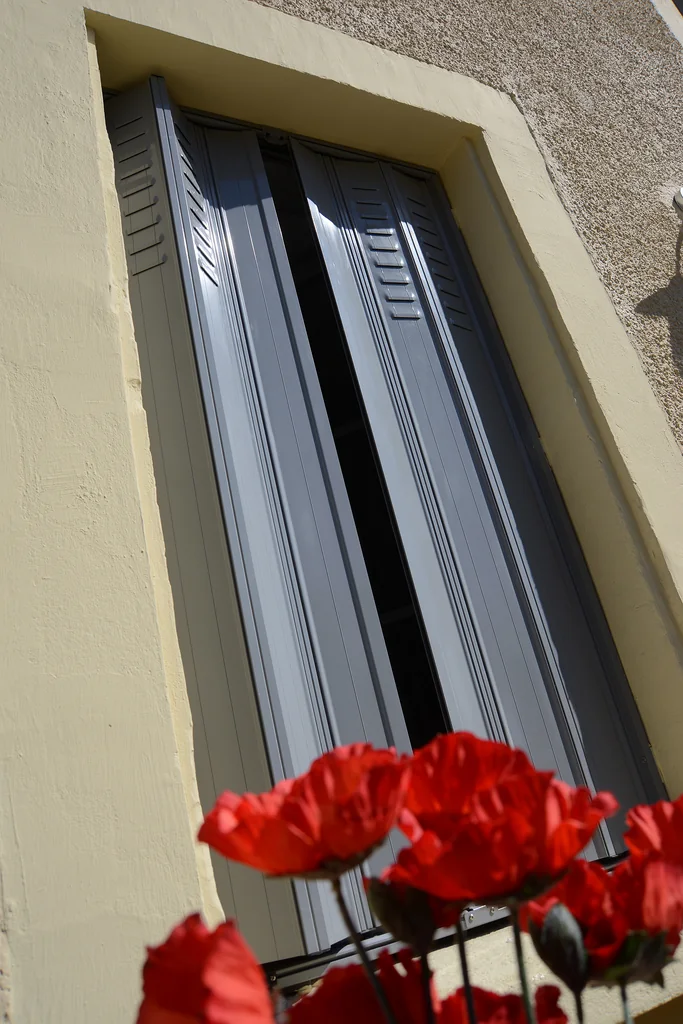 Jalousie aluminium grise sur mesure repliée dans le tableau de la fenêtre, coquelicots au premier plan