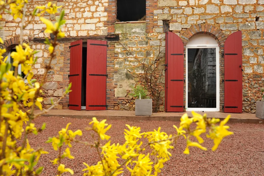 Volets battants PVC rouge basque sur mesure dont une paire cintrée, façade en pierre et briques, forsythia en fleurs